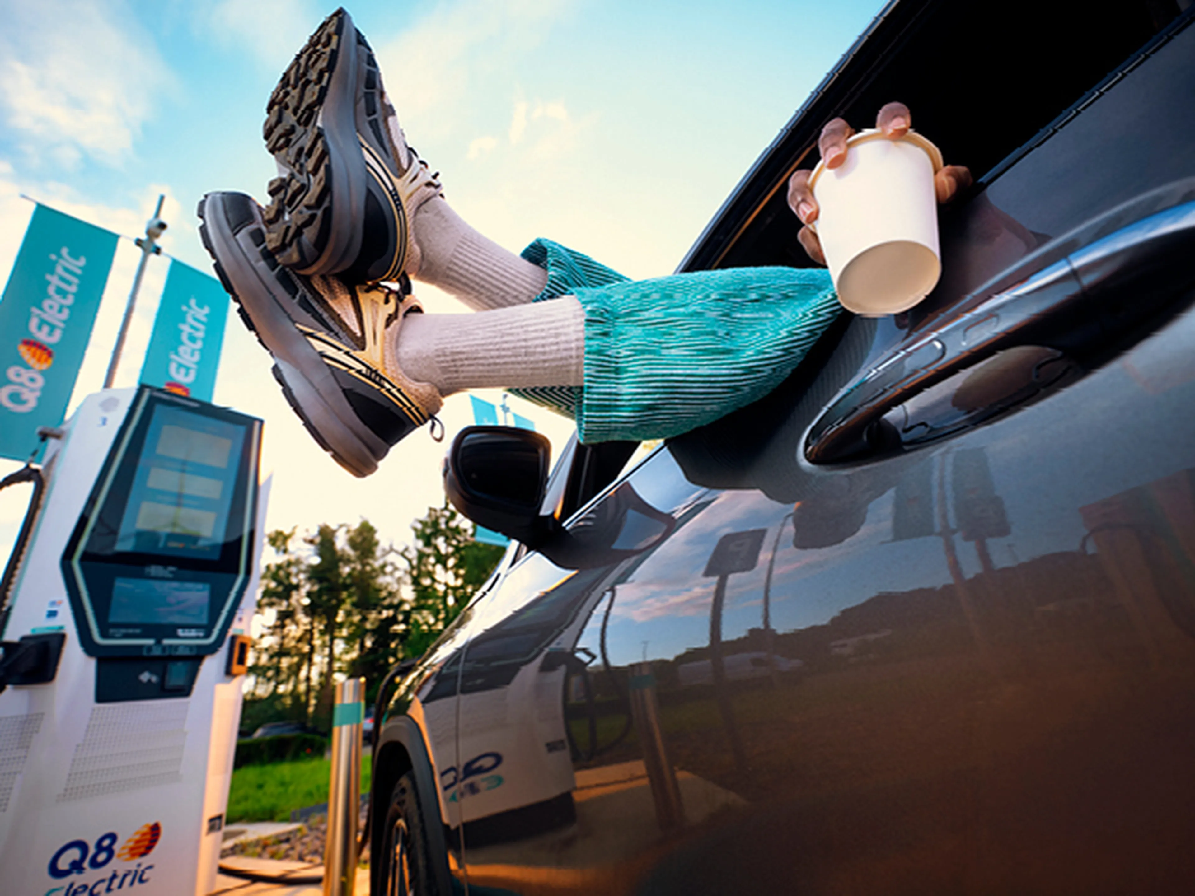 Een persoon ontspant zich in een geparkeerde auto bij een Q8 electric laadstation, met de voeten uit het raam en een koffiebeker in de hand. Op de achtergrond zijn Q8 electric vlaggen en een snellader zichtbaar.