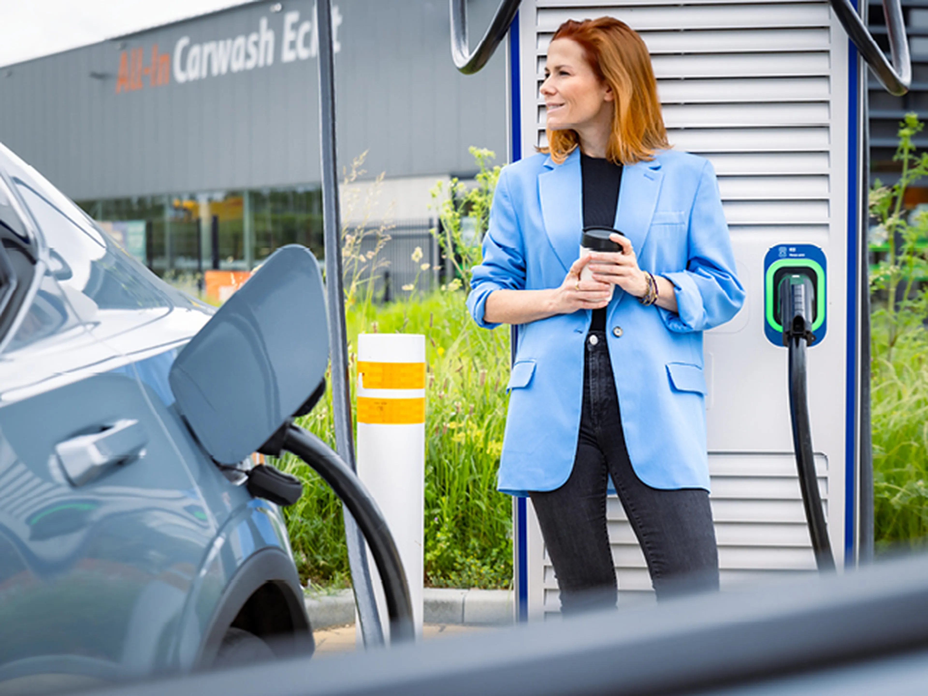 Woman waiting with a coffee next to an electric car charging at a charging station.