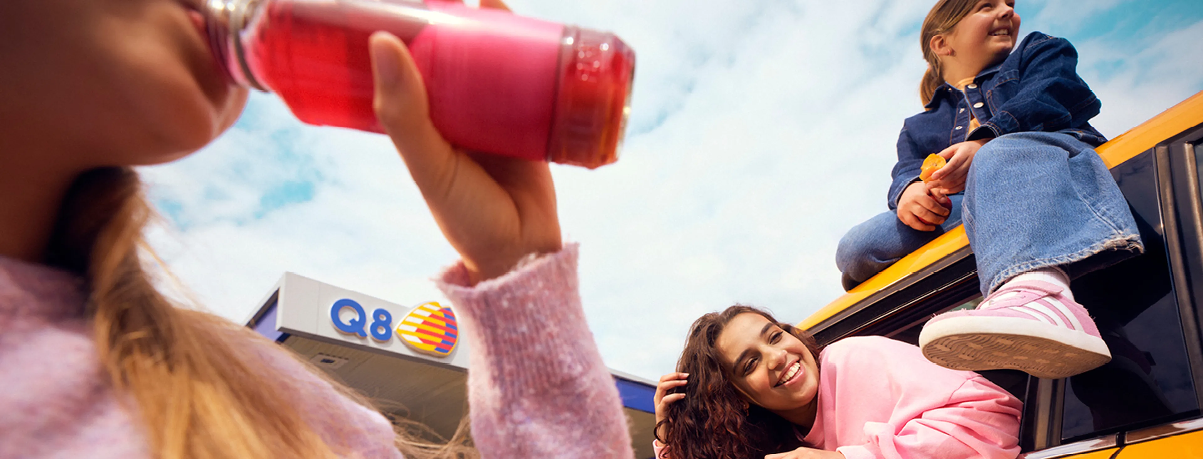 Vriendengroep neemt een pauze bij een Q8 station, terwijl iemand een drankje drinkt en anderen bij de auto ontspannen.